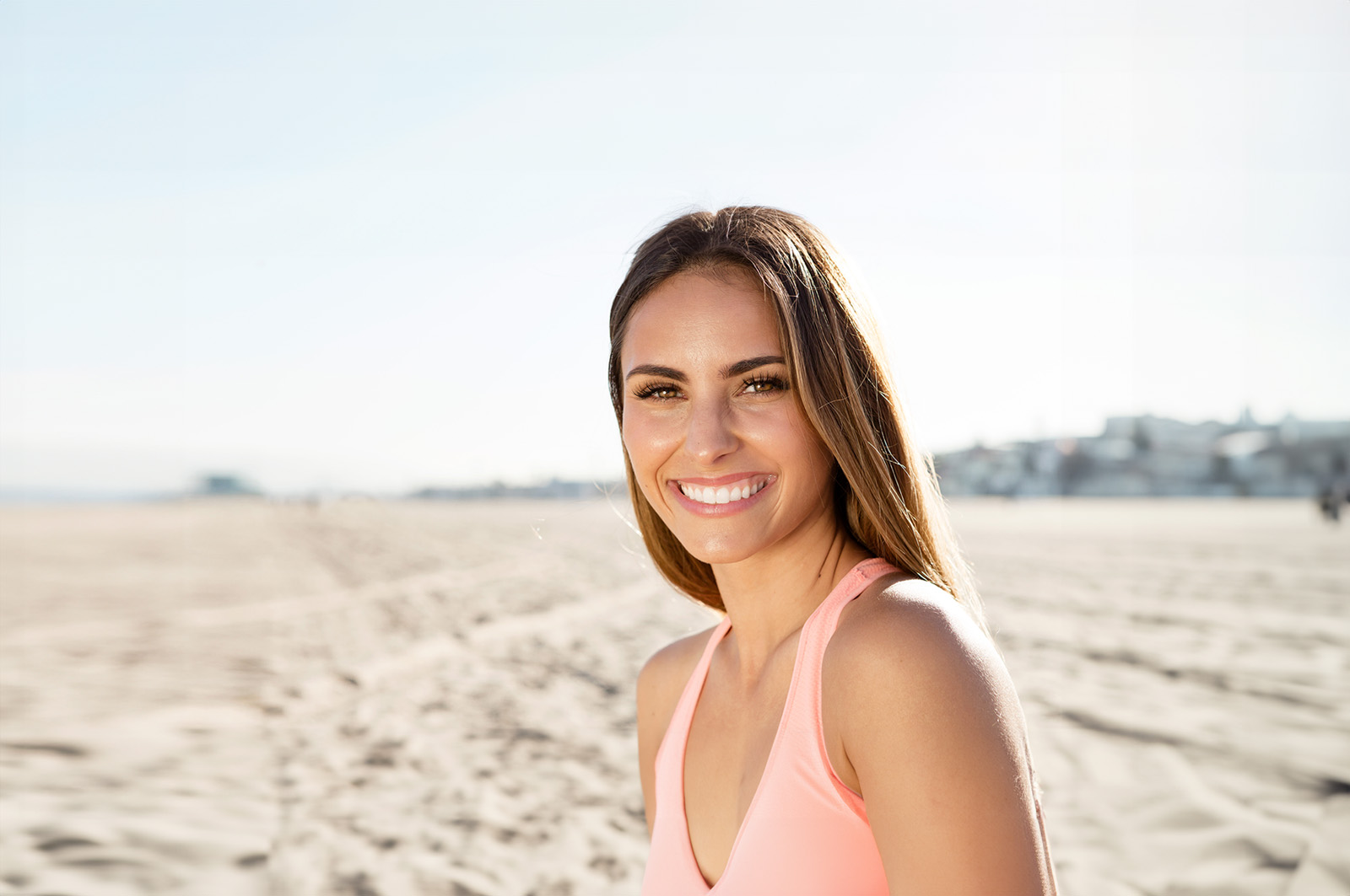 A woman with a smile stands on a sandy beach against a clear sky, wearing a light pink tank top and shorts.