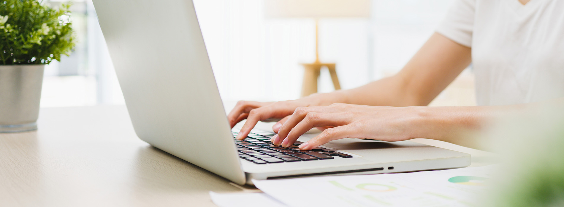This is a photograph of a person working on a laptop at a desk with a keyboard in front of them, taken from an overhead angle, showcasing a blurred background that suggests a professional environment.