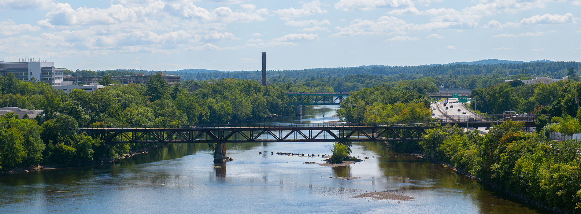 The image shows a scenic view of a river with a bridge spanning across it, surrounded by lush greenery under a clear blue sky.